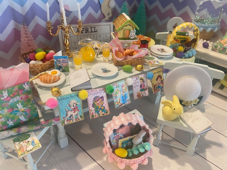 May include: A miniature Easter table setting with a white tablecloth, a gold candlestick, a pitcher of orange juice, a basket of croissants, and a variety of Easter-themed decorations, including a bunny rabbit, a basket of eggs, and a sign that says "April".