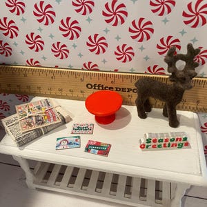Pode incluir: Uma mesa branca em miniatura com um tampo de mesa vermelho em miniatura, uma pilha de jornais em miniatura, uma figura de rena em miniatura e quatro etiquetas de presente em miniatura com tema natalino. A mesa est&aacute; sobre um fundo de papel com padr&atilde;o de bengala de caramelo vermelho e branco.