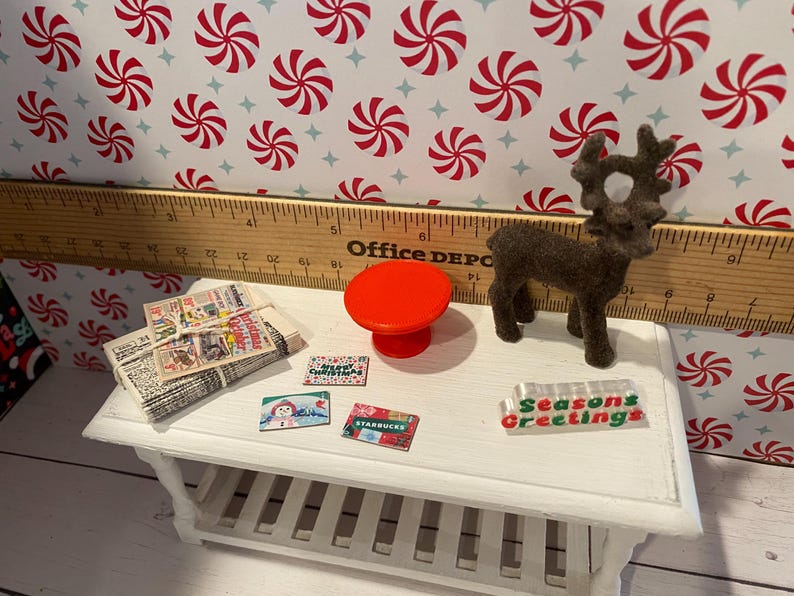 Pode incluir: Uma mesa branca em miniatura com um tampo de mesa vermelho em miniatura, uma pilha de jornais em miniatura, uma figura de rena em miniatura e quatro etiquetas de presente em miniatura com tema natalino. A mesa est&aacute; sobre um fundo de papel com padr&atilde;o de bengala de caramelo vermelho e branco.