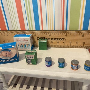 May include: Miniature baby care products including Pampers diapers, Gerber Rice cereal, and baby food jars. The items are arranged on a small white table, with a ruler and colorful striped background.