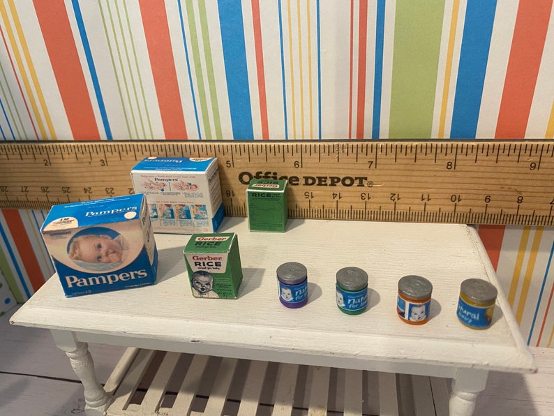 May include: Miniature baby care products including Pampers diapers, Gerber Rice cereal, and baby food jars. The items are arranged on a small white table, with a ruler and colorful striped background.