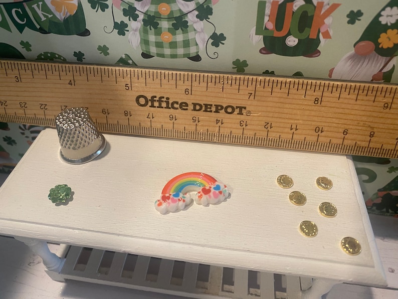 Puede incluir: Una mesa en miniatura blanca con un arco&iacute;ris, un tr&eacute;bol y monedas de oro. La mesa tiene un estante debajo. Una regla con el texto "Office Depot" est&aacute; encima de la mesa.