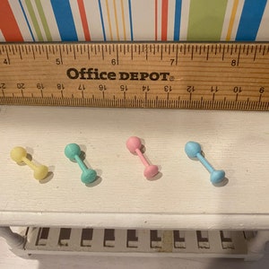 May include: Four pastel-colored miniature dumbbell toys. The dumbbells are yellow, teal, pink, and blue. A wooden ruler with the text "Office DEPOT" is above the dumbbells. The dumbbells are on a white surface.