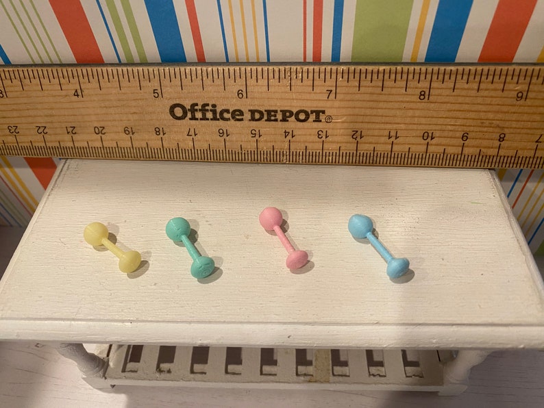 May include: Four pastel-colored miniature dumbbell toys. The dumbbells are yellow, teal, pink, and blue. A wooden ruler with the text "Office DEPOT" is above the dumbbells. The dumbbells are on a white surface.