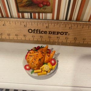 May include: A miniature roasted chicken on a white plate, garnished with red berries, tomatoes, and lemon slices. The chicken is golden brown, and the plate is set on a white surface. A wooden ruler is in the background.