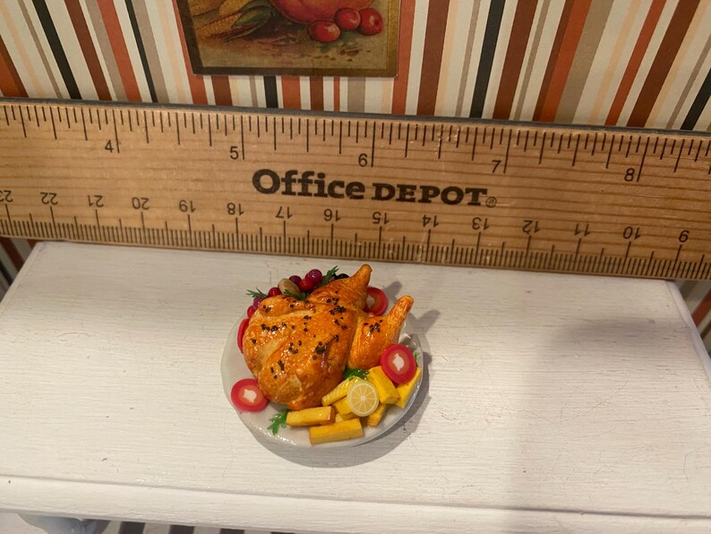 May include: A miniature roasted chicken on a white plate, garnished with red berries, tomatoes, and lemon slices. The chicken is golden brown, and the plate is set on a white surface. A wooden ruler is in the background.