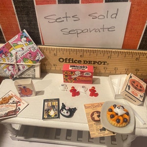 May include: A miniature Halloween scene with a variety of items, including a box of "Costume Collection" candy, a ghost-shaped pie, a book titled "It's Halloween", a box of "Candles" with a jack-o-lantern design, and a set of miniature ghost and pumpkin decorations. The text "Sets Sold Separate" is written on a piece of paper in the background.