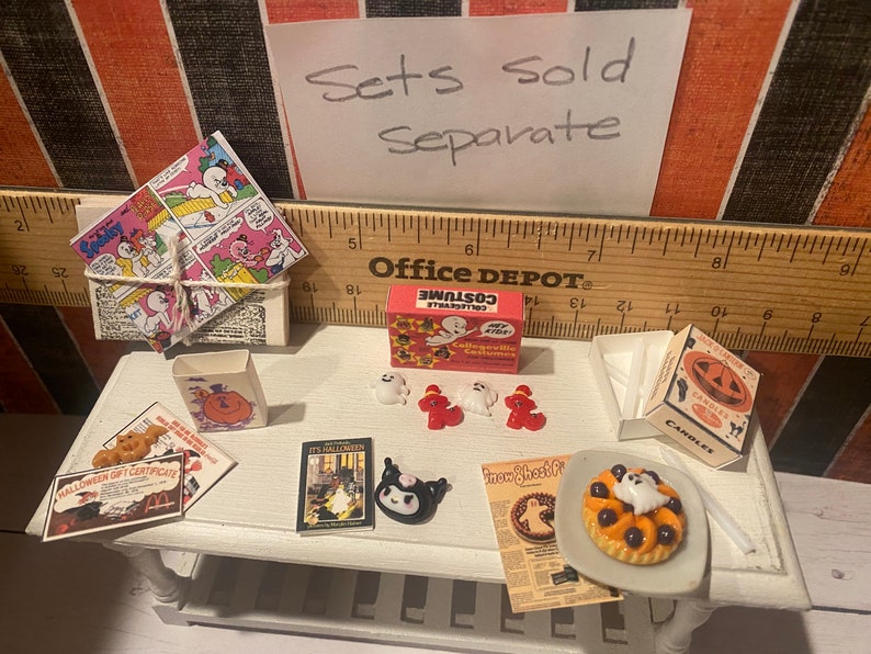 May include: A miniature Halloween scene with a variety of items, including a box of "Costume Collection" candy, a ghost-shaped pie, a book titled "It's Halloween", a box of "Candles" with a jack-o-lantern design, and a set of miniature ghost and pumpkin decorations. The text "Sets Sold Separate" is written on a piece of paper in the background.