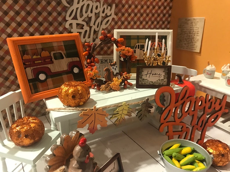 May include: A miniature Thanksgiving table setting with a white table, two white chairs, a red truck with pumpkins, a fall wreath, a sign that says "Home", and a sign that says "Happy Fall".