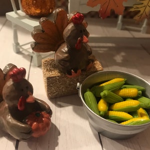 May include: Two brown ceramic turkey figurines, one holding a small orange pumpkin, the other sitting on a bale of hay. A small metal bucket filled with miniature corn on the cob.