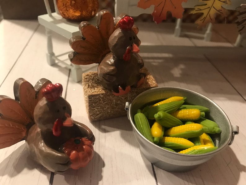 May include: Two brown ceramic turkey figurines, one holding a small orange pumpkin, the other sitting on a bale of hay. A small metal bucket filled with miniature corn on the cob.