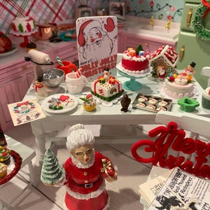 May include: A miniature dollhouse kitchen scene decorated for Christmas. The scene includes a table with a variety of miniature Christmas treats, including cakes, cookies, and a gingerbread house. There is also a miniature Santa Claus figurine holding a Christmas tree. The text "Merry Christmas" is displayed on a red felt banner.