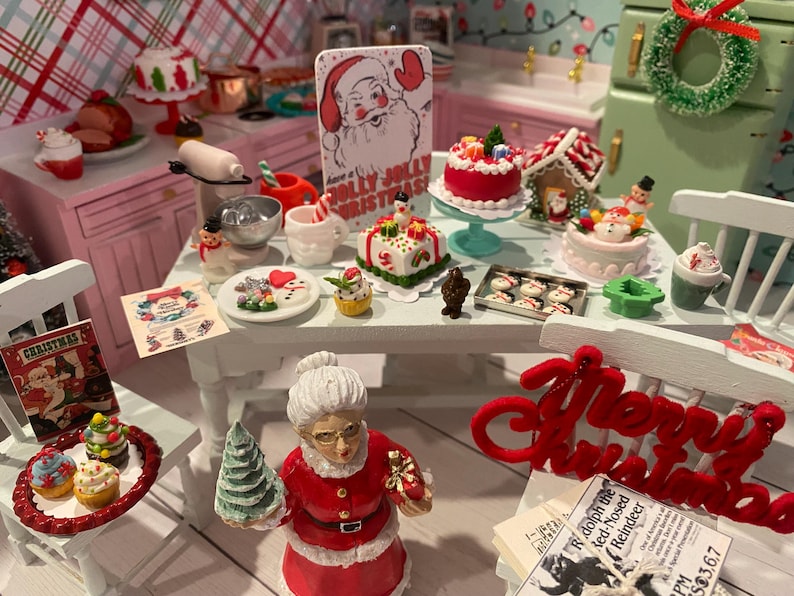 May include: A miniature dollhouse kitchen scene decorated for Christmas. The scene includes a table with a variety of miniature Christmas treats, including cakes, cookies, and a gingerbread house. There is also a miniature Santa Claus figurine holding a Christmas tree. The text "Merry Christmas" is displayed on a red felt banner.