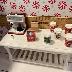 Pode incluir: Uma mesa de caf&eacute; branca com uma prateleira, uma cafeteira vermelha, uma caixa de caf&eacute; Starbucks Holiday Blend e cinco x&iacute;caras de caf&eacute; de tema natalino vermelhas e verdes.