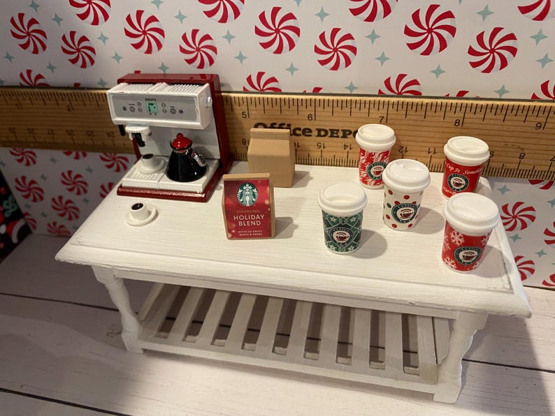 Pode incluir: Uma mesa de caf&eacute; branca com uma prateleira, uma cafeteira vermelha, uma caixa de caf&eacute; Starbucks Holiday Blend e cinco x&iacute;caras de caf&eacute; de tema natalino vermelhas e verdes.