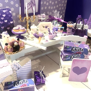 May include: A miniature purple and white themed party scene with a unicorn, cupcakes, and a table set with purple and white dishes, a purple and white striped candy cane, and two boxes of Fanta grape soda.