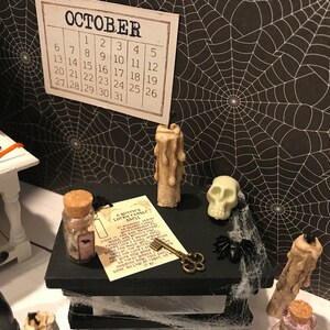 Puede incluir: Una escena de Halloween en miniatura con un fondo de telara&ntilde;a blanco y negro. Una peque&ntilde;a caja de madera con tapa negra est&aacute; decorada con una calavera blanca, una peque&ntilde;a botella con tap&oacute;n de corcho, una llave, un trozo de papel con texto y dos peque&ntilde;as velas. El texto en el papel dice "A Witch's Lucky Candle Spell".