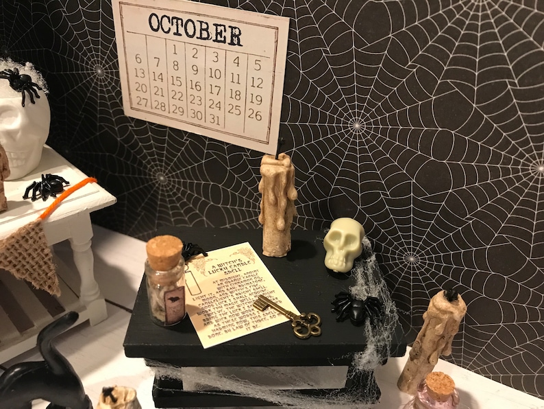 Puede incluir: Una escena de Halloween en miniatura con un fondo de telara&ntilde;a blanco y negro. Una peque&ntilde;a caja de madera con tapa negra est&aacute; decorada con una calavera blanca, una peque&ntilde;a botella con tap&oacute;n de corcho, una llave, un trozo de papel con texto y dos peque&ntilde;as velas. El texto en el papel dice "A Witch's Lucky Candle Spell".