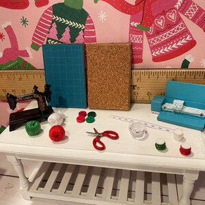 May include: Miniature craft supplies on a white table. Includes a black sewing machine, blue cutting mat, cork board, red scissors, measuring tape, and spools of thread in various colors. A blue cutting machine is also present.