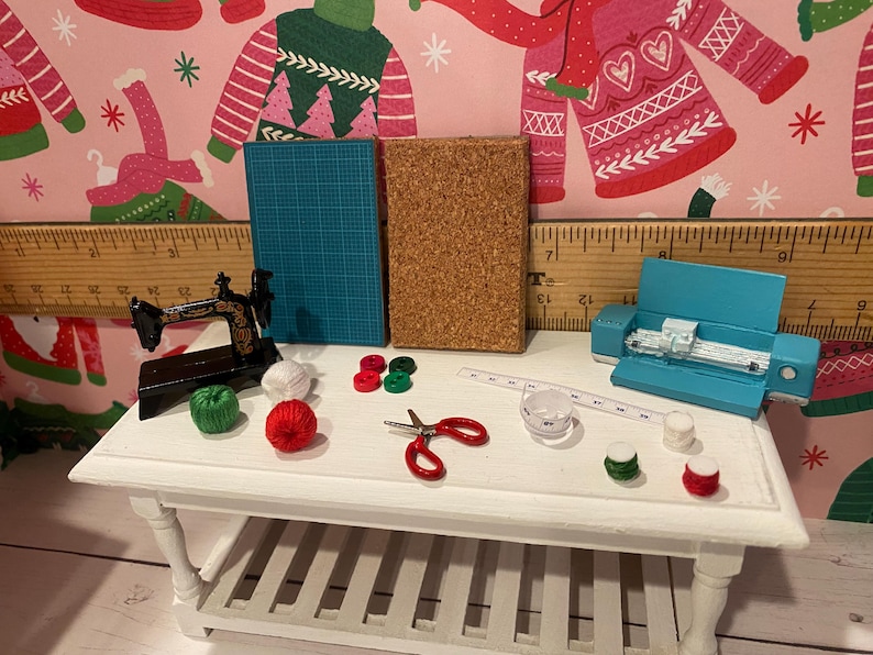May include: Miniature craft supplies on a white table. Includes a black sewing machine, blue cutting mat, cork board, red scissors, measuring tape, and spools of thread in various colors. A blue cutting machine is also present.