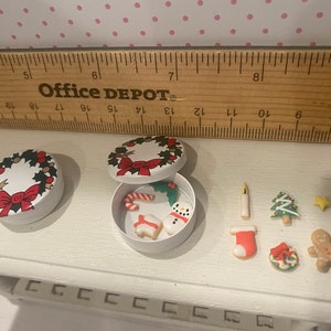 May include: Two white round tins with a Christmas wreath design. One tin is open and contains small Christmas-themed cookies, including a snowman, candy cane, and a gingerbread house. The other tin is closed. The cookies are arranged on a white surface.