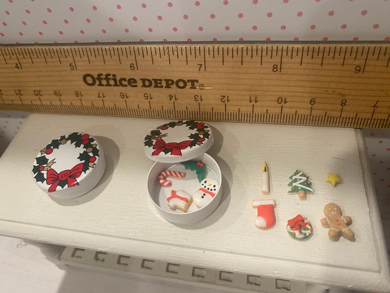 May include: Two white round tins with a Christmas wreath design. One tin is open and contains small Christmas-themed cookies, including a snowman, candy cane, and a gingerbread house. The other tin is closed. The cookies are arranged on a white surface.