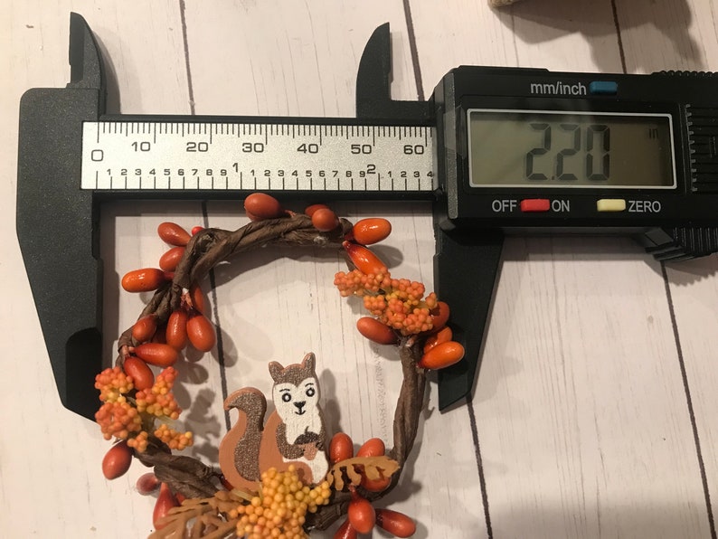 May include: A brown twig wreath with orange berry accents and a small wooden squirrel figurine. The wreath measures 220 mm in diameter.