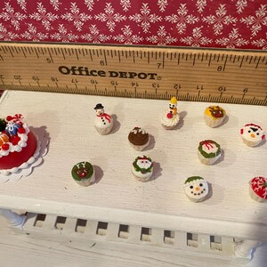 May include: Miniature Christmas-themed desserts on a white surface. The collection includes a red cake with gifts, snowmen, and cupcakes decorated with festive designs. A wooden ruler is in the background.