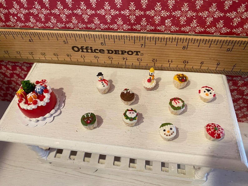 May include: Miniature Christmas-themed desserts on a white surface. The collection includes a red cake with gifts, snowmen, and cupcakes decorated with festive designs. A wooden ruler is in the background.