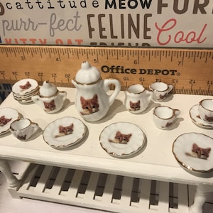 May include: A miniature tea set with a cat theme. The set includes a teapot, creamer, sugar bowl, four teacups, and four saucers. The pieces are white with gold trim and feature a brown tabby cat design.