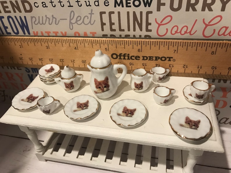 May include: A miniature tea set with a cat theme. The set includes a teapot, creamer, sugar bowl, four teacups, and four saucers. The pieces are white with gold trim and feature a brown tabby cat design.