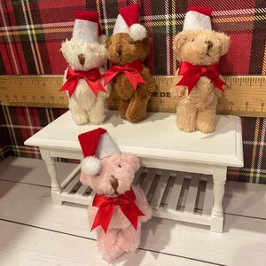 Puede incluir: Cuatro osos de peluche con gorros de Pap&aacute; Noel y lazos rojos. Los osos son de varios colores: blanco, marr&oacute;n y rosa. Est&aacute;n colocados sobre una peque&ntilde;a mesa blanca con una regla de madera en el fondo. El fondo es un estampado de cuadros rojos y verdes.