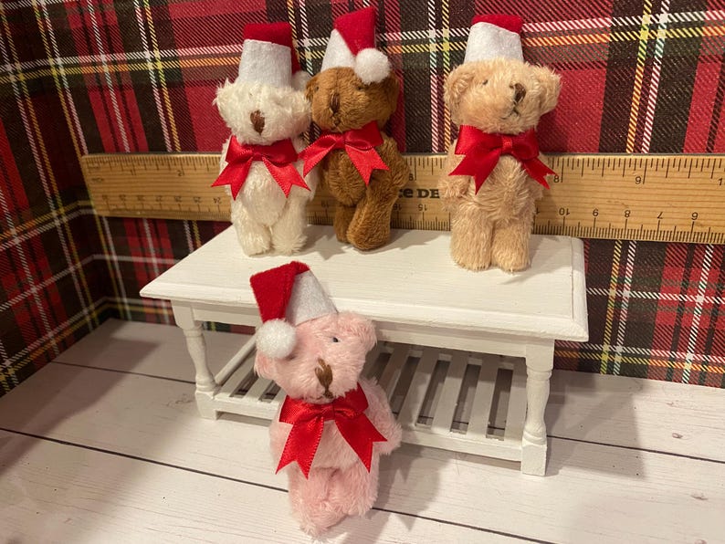 Puede incluir: Cuatro osos de peluche con gorros de Pap&aacute; Noel y lazos rojos. Los osos son de varios colores: blanco, marr&oacute;n y rosa. Est&aacute;n colocados sobre una peque&ntilde;a mesa blanca con una regla de madera en el fondo. El fondo es un estampado de cuadros rojos y verdes.