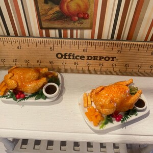May include: Two miniature roasted chicken dishes on white plates. Each plate includes a golden-brown chicken, french fries, vegetables, and a small bowl of gravy. The dishes are set on a white surface.