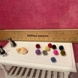 K&ouml;nnte beinhalten: Zwei Miniatur-Katzenfiguren, eine schwarz-wei&szlig;e und eine graue, neben einer kleinen orangefarbenen Katzenfigur und zehn winzigen, bunten Filzkugeln. Ein Holzlineal befindet sich hinter den Objekten und zeigt eine Skala von etwa 20 cm.