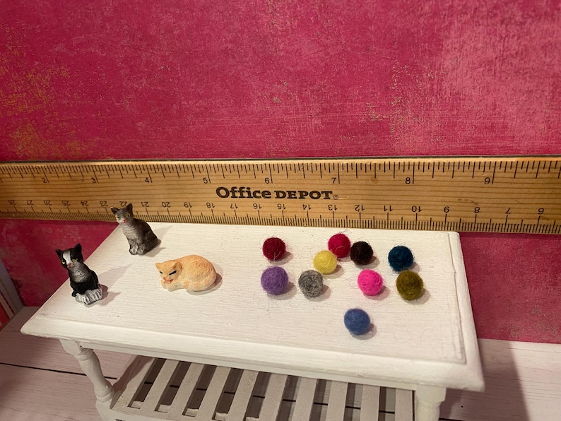 K&ouml;nnte beinhalten: Zwei Miniatur-Katzenfiguren, eine schwarz-wei&szlig;e und eine graue, neben einer kleinen orangefarbenen Katzenfigur und zehn winzigen, bunten Filzkugeln. Ein Holzlineal befindet sich hinter den Objekten und zeigt eine Skala von etwa 20 cm.
