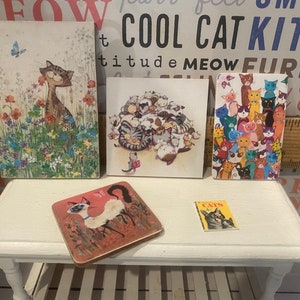 May include: Five miniature framed pictures of cats, with a small book titled "Cats" in front of them. The frames are white and the pictures are colorful. The book is red, white, and yellow.