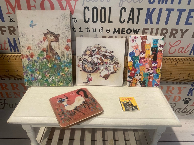 May include: Five miniature framed pictures of cats, with a small book titled "Cats" in front of them. The frames are white and the pictures are colorful. The book is red, white, and yellow.
