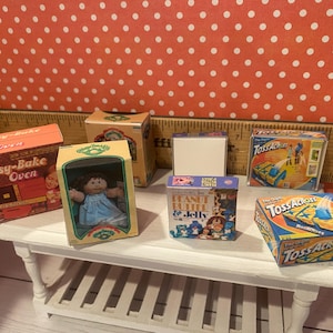 Op de afbeelding: Een verzameling miniatuur speelgoeddozen en een pop zijn gerangschikt op een witte tafel. De dozen bevatten een "Easy-Bake Oven", "Peanut Butter & Jelly" en het spel "Toss Across". Een Cabbage Patch Kids pop zit in een doos.