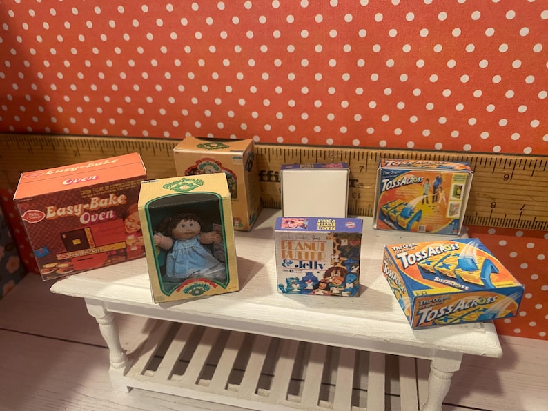 Op de afbeelding: Een verzameling miniatuur speelgoeddozen en een pop zijn gerangschikt op een witte tafel. De dozen bevatten een "Easy-Bake Oven", "Peanut Butter & Jelly" en het spel "Toss Across". Een Cabbage Patch Kids pop zit in een doos.
