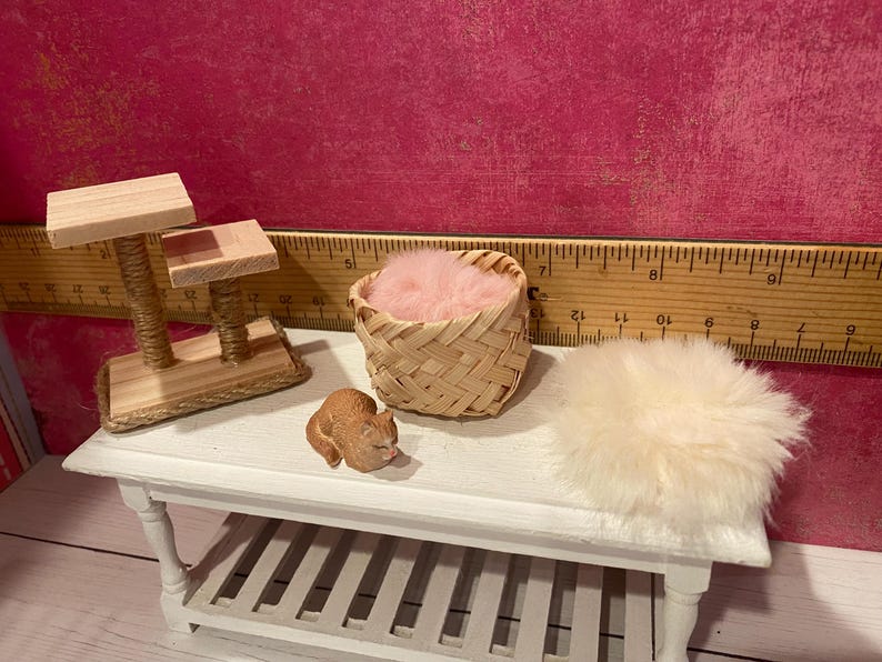 K&ouml;nnte beinhalten: Ein Miniatur-Kratzbaum aus Holz mit zwei Plattformen, ein kleiner geflochtener Korb mit rosa Kunstfell, eine winzige braune Katzenfigur und ein runder wei&szlig;er Kunstfellteppich. Ein Holzlineal im Hintergrund zeigt eine Skala von 30 Zentimetern.