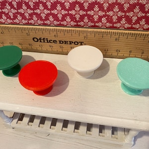 May include: Four colorful cabinet knobs in green, red, white, and teal are displayed on a white table. The knobs have a round top and a short, pedestal base. A wooden ruler with the Office Depot logo is in the background.