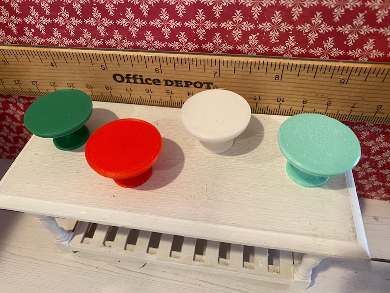 May include: Four colorful cabinet knobs in green, red, white, and teal are displayed on a white table. The knobs have a round top and a short, pedestal base. A wooden ruler with the Office Depot logo is in the background.