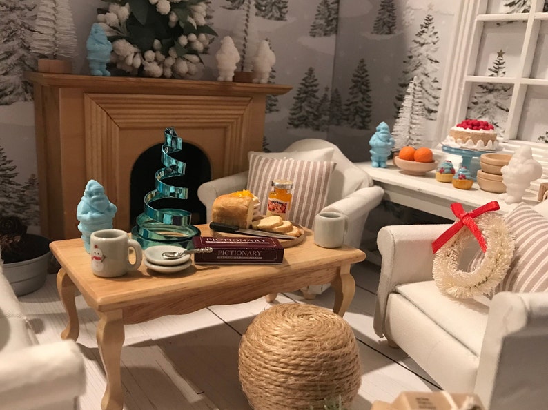 May include: A miniature living room scene with white furniture, a fireplace, a coffee table with a game of Pictionary, a blue spiral Christmas tree, and a wreath with a red bow.