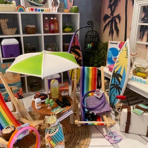Peut inclure: Une scène de plage miniature comprenant deux chaises longues en bois avec un tissu rayé arc-en-ciel, un parasol vert et blanc, une table ronde en bois avec des boissons et accessoires miniatures, une planche de surf, une valise blanche et divers décors.
