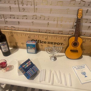 May include: A miniature Hanukkah scene with a menorah, candles, a bottle of wine, two wine glasses, a box of Chanukah candles, a songbook, and a miniature acoustic guitar.