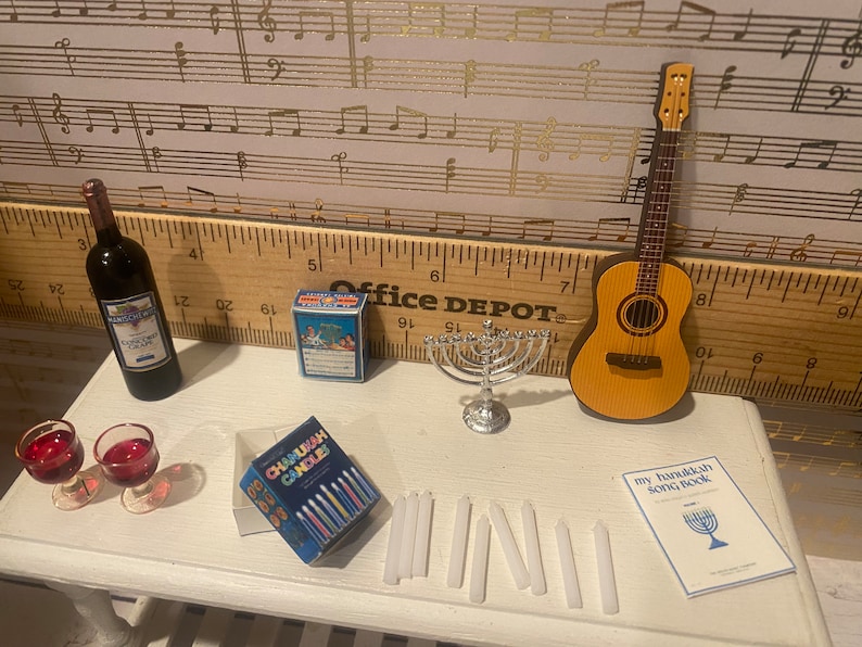 May include: A miniature Hanukkah scene with a menorah, candles, a bottle of wine, two wine glasses, a box of Chanukah candles, a songbook, and a miniature acoustic guitar.