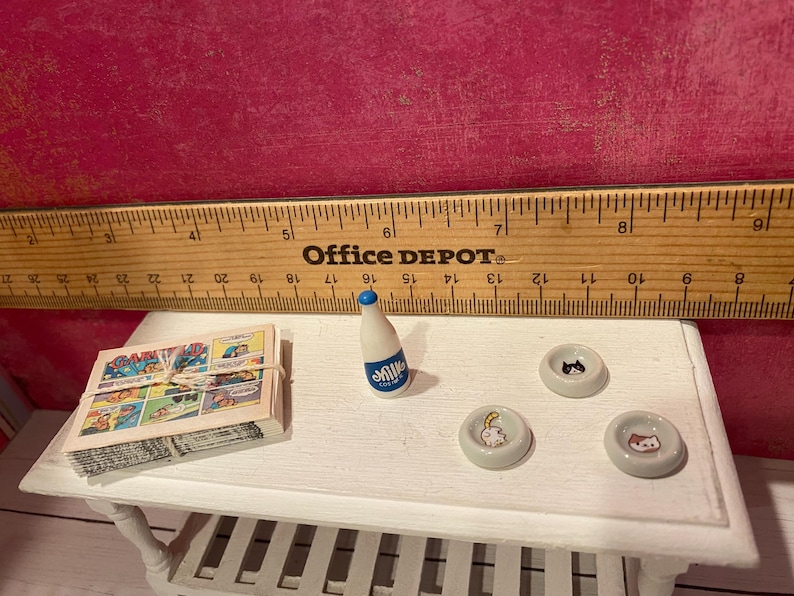 K&ouml;nnte beinhalten: Ein Miniatur-Set bestehend aus einem mit Schnur gebundenen Stapel Comics, einer kleinen wei&szlig;en Flasche mit blauem Verschluss und der Aufschrift Milk sowie drei winzigen wei&szlig;en Schalen mit Katzenmotiven, platziert neben einem Holzlineal.