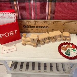 Puede incluir: Una escena navide&ntilde;a en miniatura con un buz&oacute;n rojo con la palabra "POST", un tren de madera y un plato de galletas festivas. Se ve una tarjeta de "Carta a Pap&aacute; Noel". La escena est&aacute; sobre una mesa blanca.