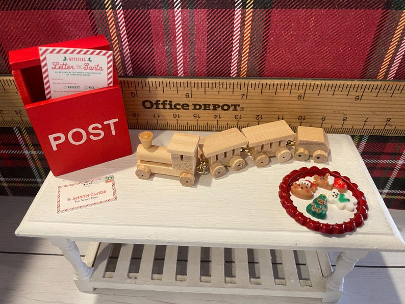 Puede incluir: Una escena navide&ntilde;a en miniatura con un buz&oacute;n rojo con la palabra "POST", un tren de madera y un plato de galletas festivas. Se ve una tarjeta de "Carta a Pap&aacute; Noel". La escena est&aacute; sobre una mesa blanca.